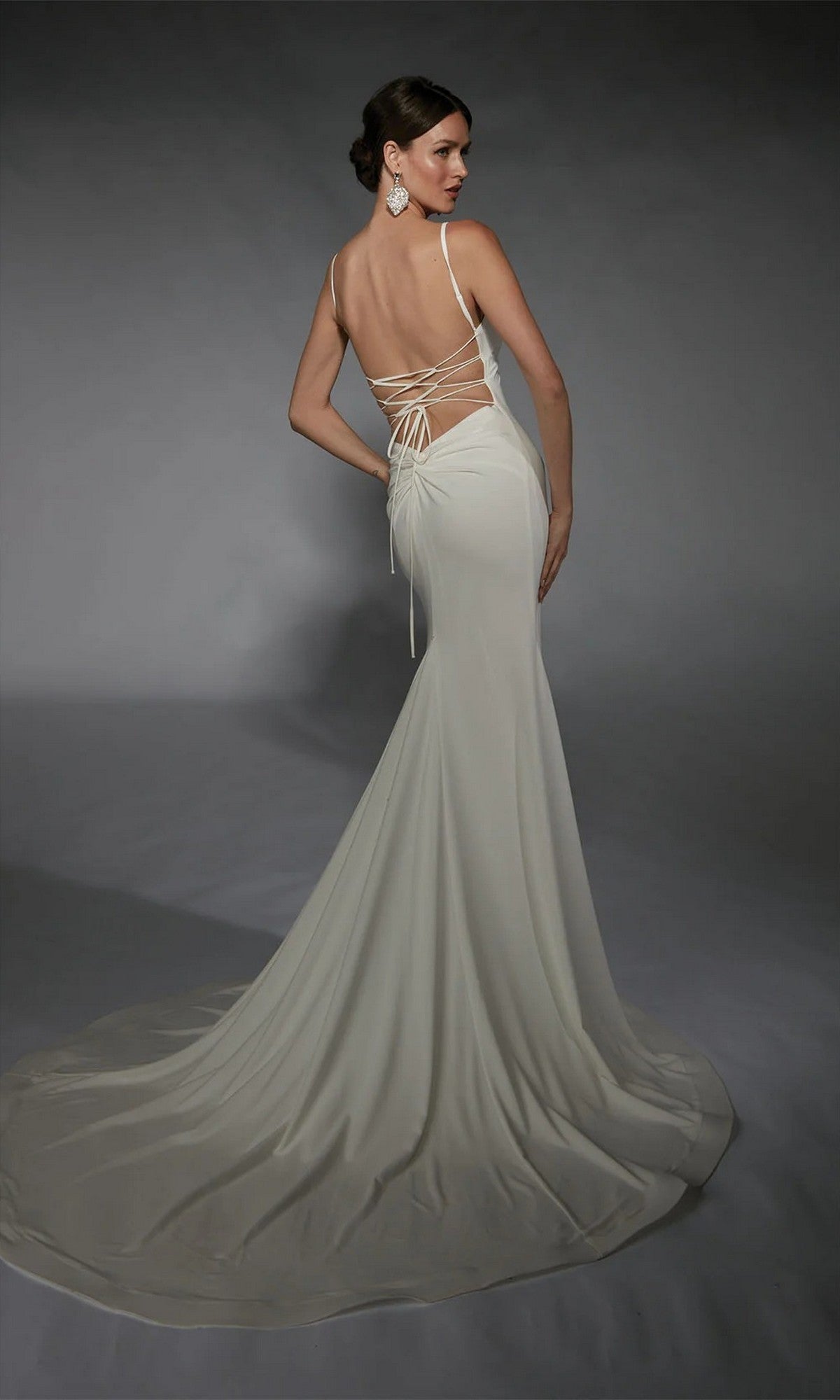A woman poses in the Alyce 7108 long white dress, a sleeveless formal gown featuring lace-up detailing, a backless design, and a train, against a gray background.